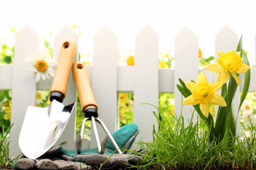 The image depicts a section of a garden with a well-maintained grassy lawn in the foreground, bordered by a small flower bed containing bright yellow daffodils and a white daisy with a yellow center. Behind the flower bed, there is a white wooden picket fence, suggesting a front or backyard setting typical of residential gardens in Willesden. The garden scene is bright and illuminated by natural sunlight, indicating a clear day with no visible weather disturbances. In front of the fence, gardening tools including a small hand trowel with a white blade, a pair of pruning shears, and a gardening glove are arranged on the ground, hinting at recent or ongoing garden maintenance activities. The plants and lawn appear healthy and lush, with the flower bed adding vibrant natural color to the scene. This setup illustrates typical outdoor garden features that could benefit from expert gardening and landscaping services by Pressure Washing Willesden, helping to preserve outdoor spaces in good condition and enhance curb appeal.