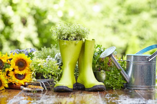 This image depicts a garden scene with a weathered wooden outdoor table displaying gardening tools and decorative elements. In the foreground, a pair of bright green rubber boots filled with small white and green flowering plants serve as a creative planter, standing upright on the table. To the left, a cluster of vibrant yellow sunflowers and smaller mixed flowers adds a splash of colour, contrasting with the natural tones of the wood and surrounding greenery. On the right side, a silver metal watering can with a long spout and a blue-handled garden trowel are positioned nearby, suggesting outdoor maintenance activities. The background features lush, blurred foliage with trees and shrubs, indicating a well-maintained garden space in a temperate environment. The scene is illuminated by natural sunlight, creating a fresh and inviting outdoor atmosphere associated with gardening and landscaping services in Willesden, London. This setup reflects the elements typically found in a nurturing garden environment, subtly aligning with gardening service offerings such as planting, lawn care, and general outdoor maintenance by Pressure Washing Willesden, adding a natural connection to professional garden care in the local area.
